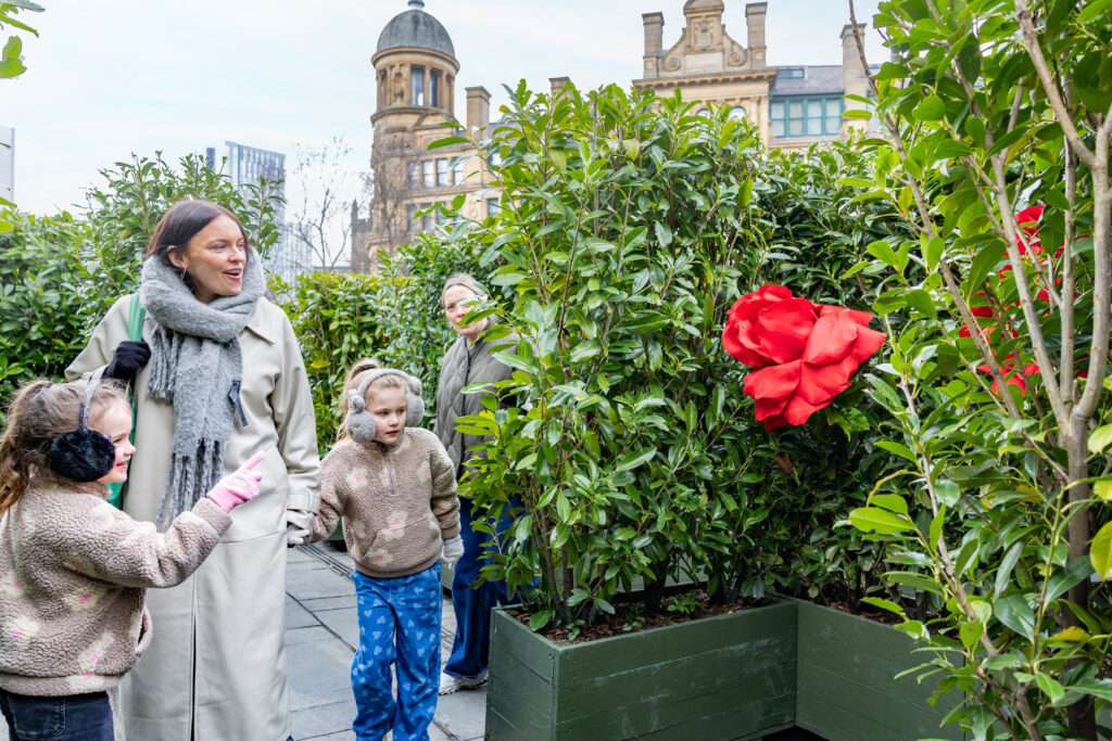 Two children and two adults exploring plants with large artificial red flowers in an urban garden