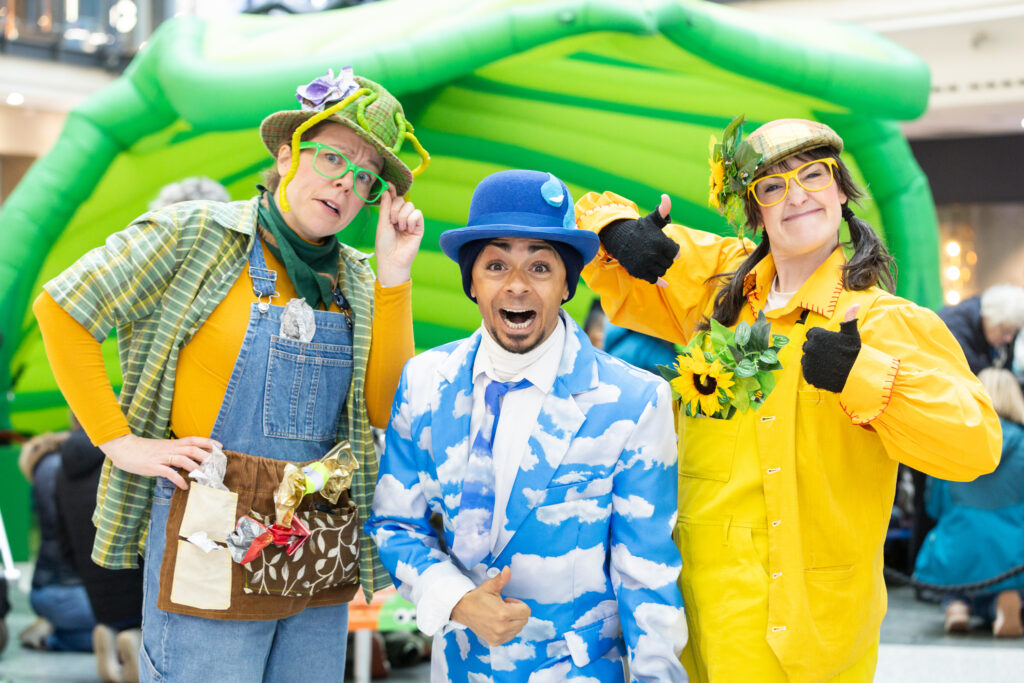 Three people in colourful costumes posing and giving thumbs up at an indoor event