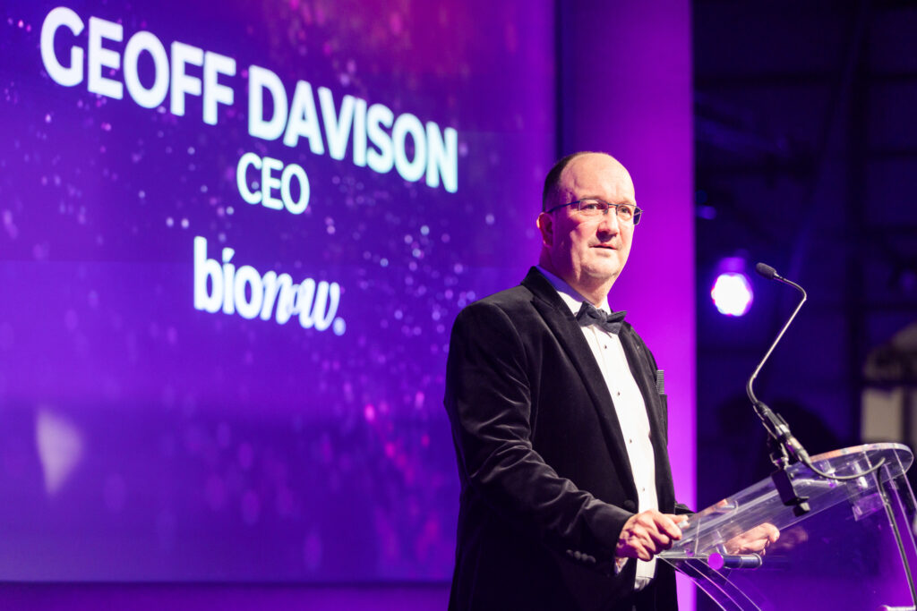 Man in tuxedo standing at a clear podium speaking on stage with purple background