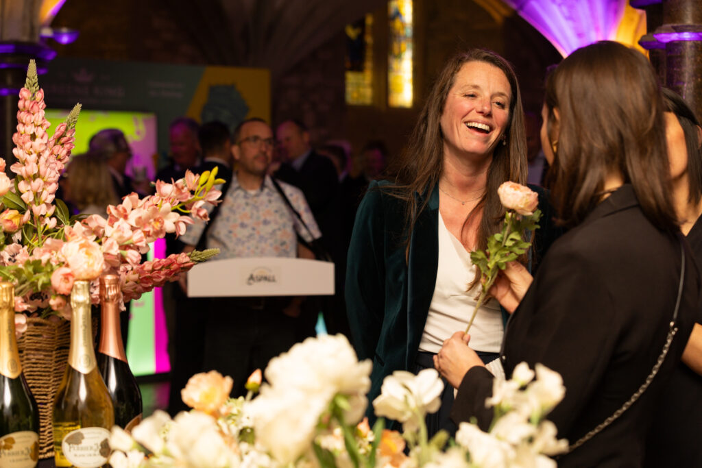Two women talking and smiling near floral arrangements and bottles at an indoor event