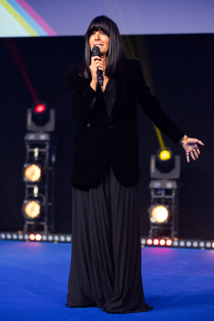 Woman with long dark hair speaking into microphone on stage with coloured lights