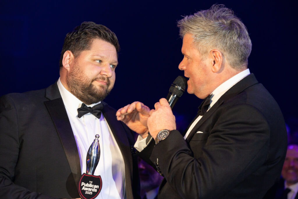 Two men in tuxedos at awards event, one holding The Publican Awards 2025 trophy, other speaking into microphone