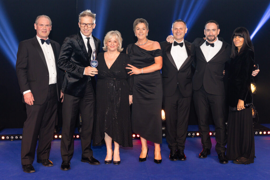 Group of six people in formalwear standing on stage with blue lighting, one holding an award