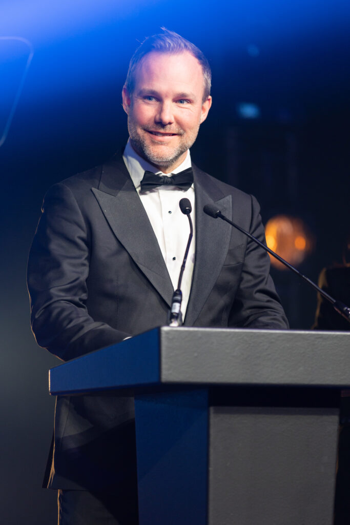 Man in tuxedo speaking at podium with microphone in dimly lit event space