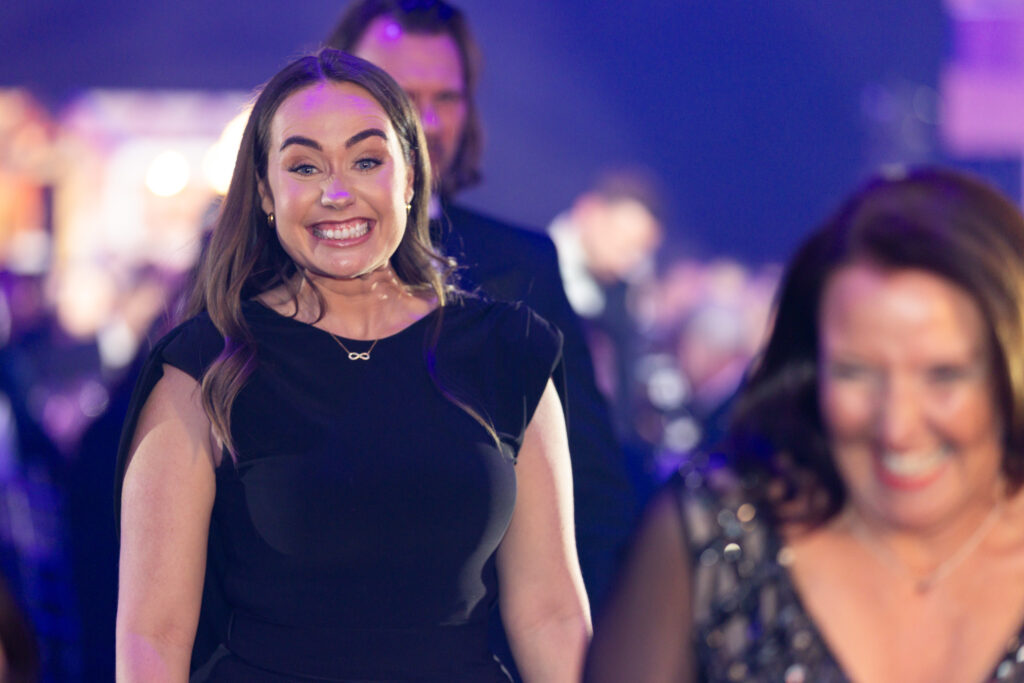 Woman in black dress smiling widely at an indoor event with blurred crowd behind