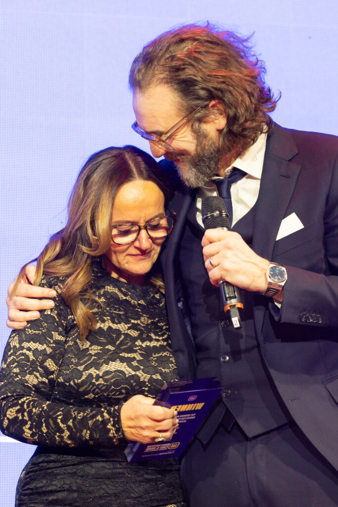 Man in suit holding microphone embraces woman in black lace dress holding award plaque