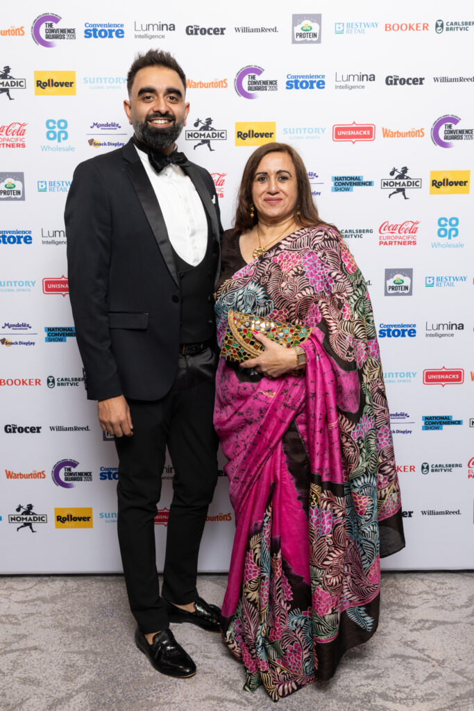 Man in black tuxedo and woman in floral pink saree posing against sponsor backdrop