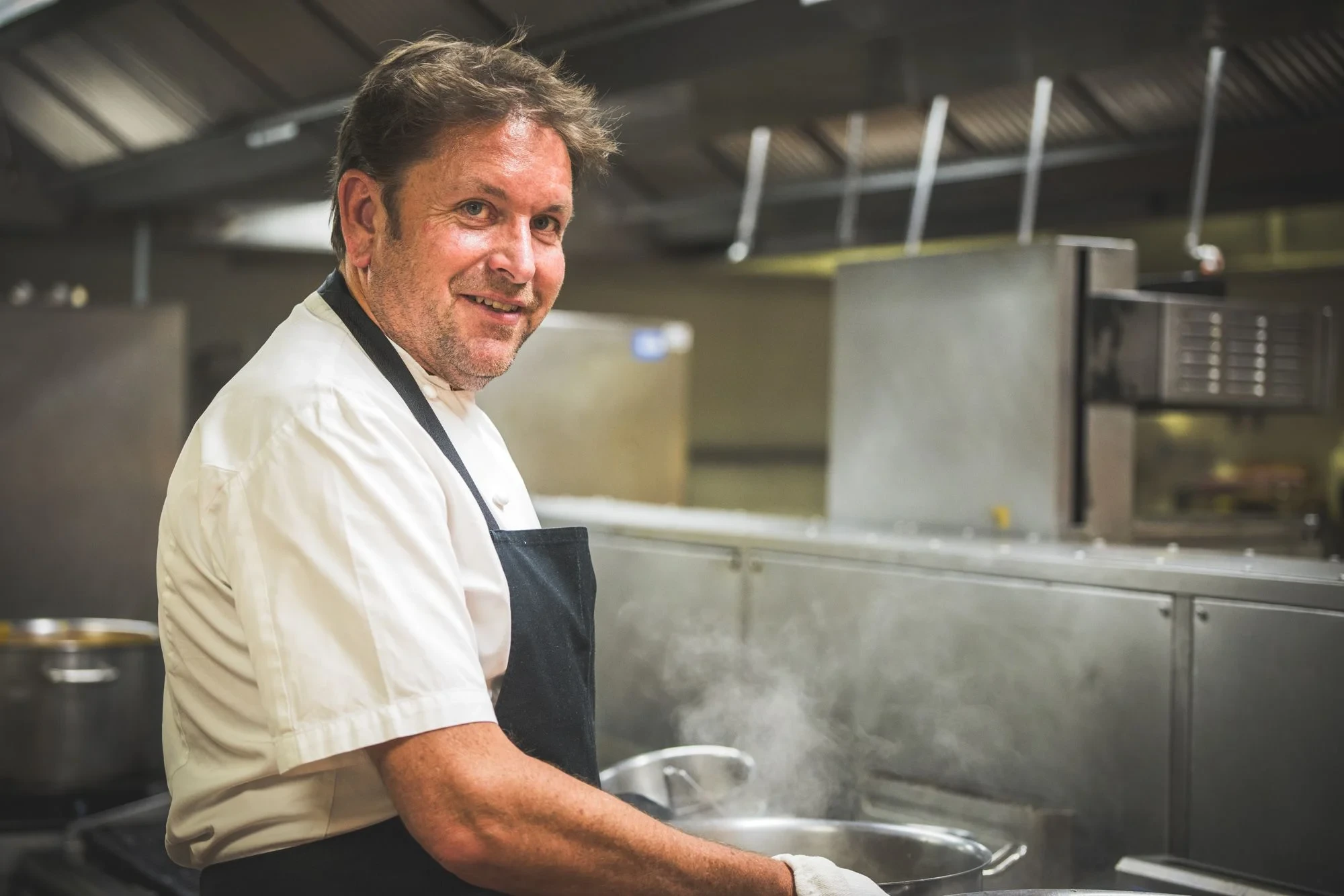 James Martin wearing a white chef jacket and black apron stands at a stove with a steaming pan.