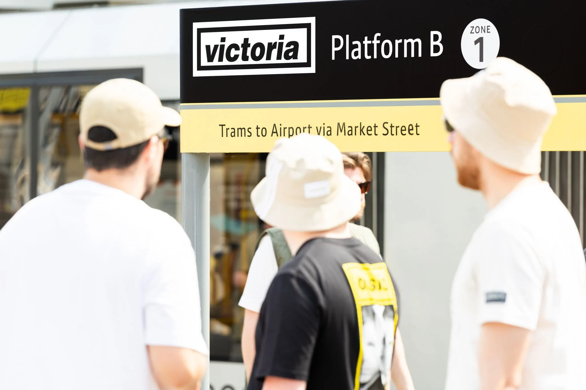 Four people stand near a Victoria Station Platform B sign, with two men wearing cream hats facing away.