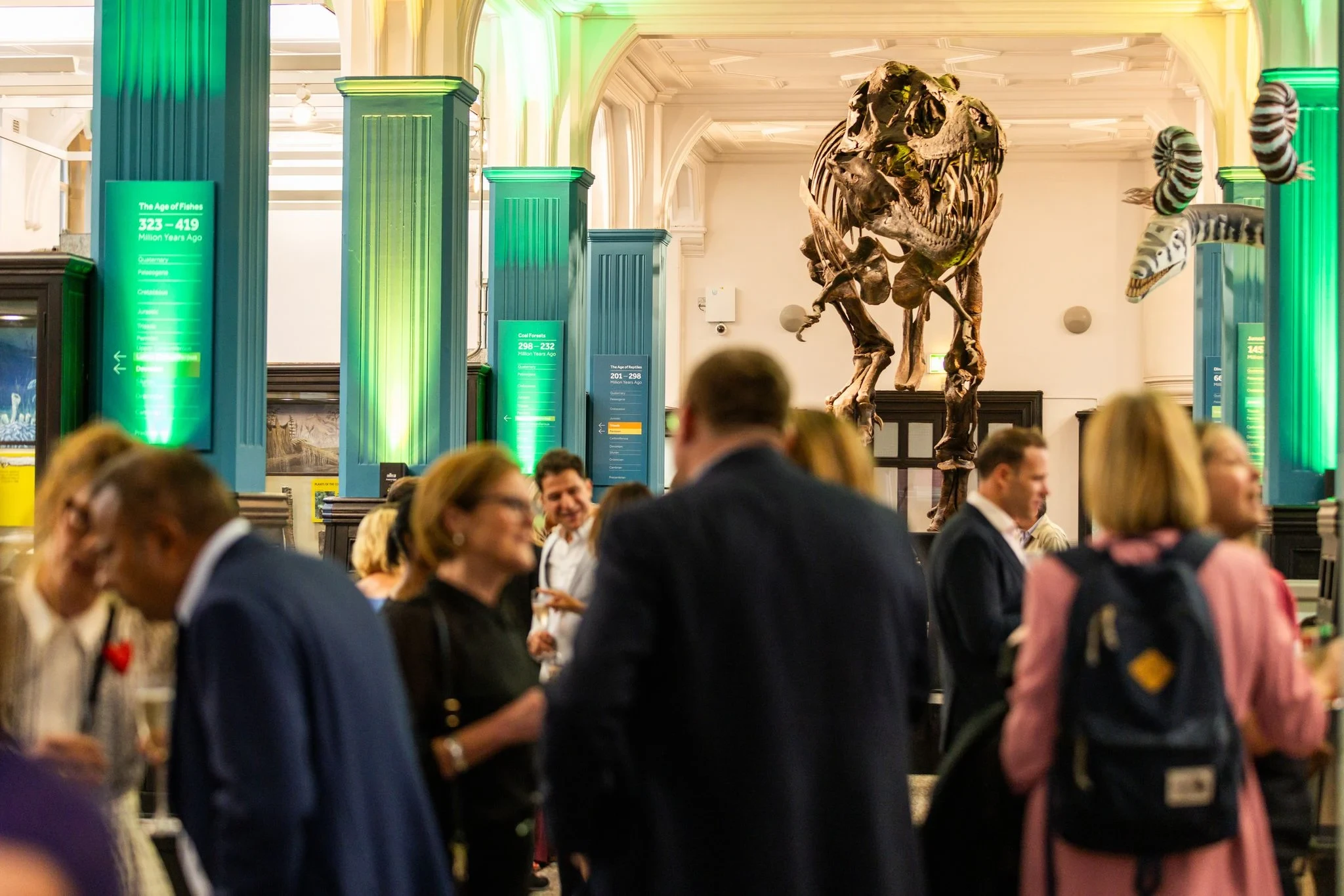 Group of people standing and talking inside a hall with dinosaur skeleton and lit columns.