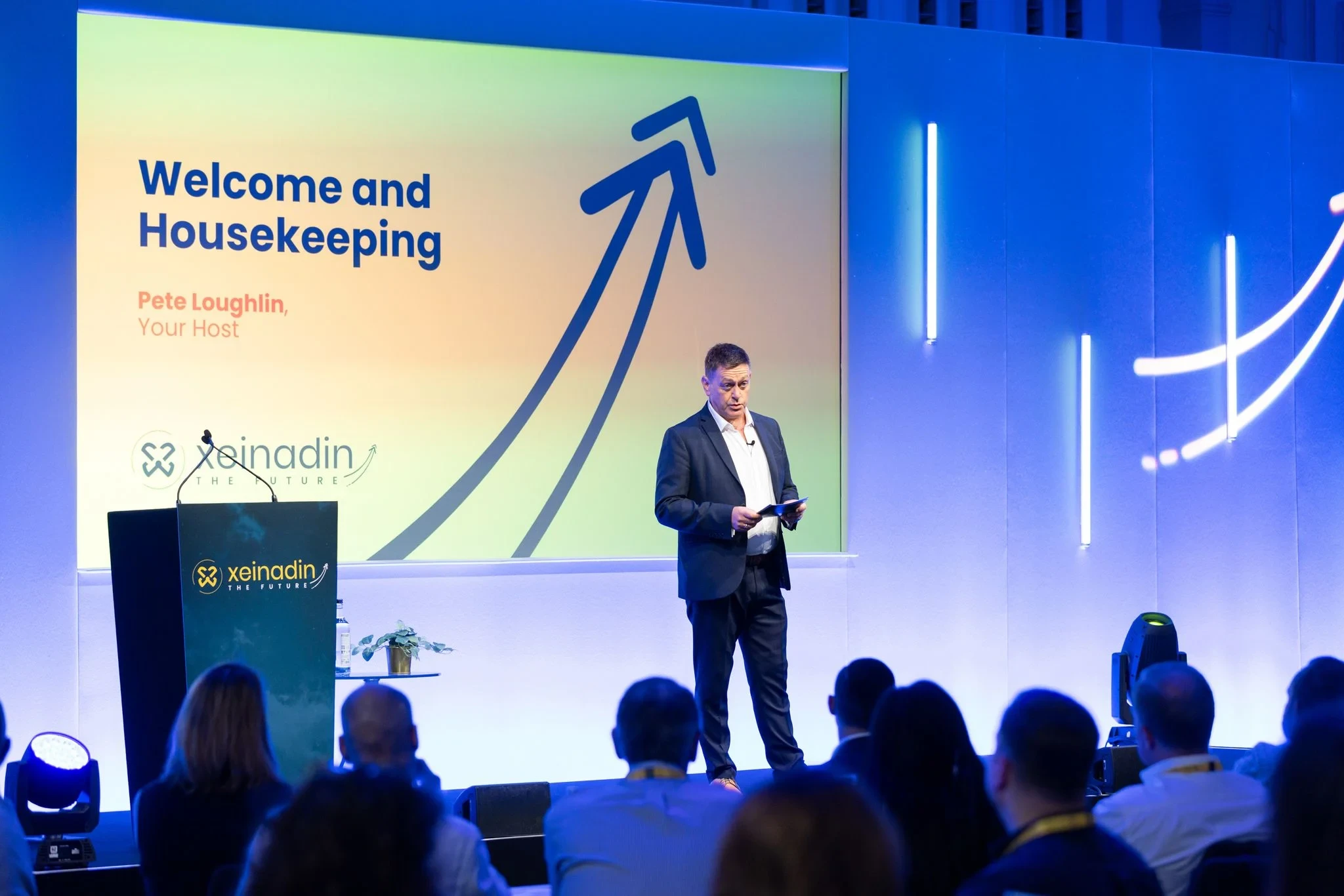 Man in dark suit stands on stage speaking to seated audience under a screen displaying 'Welcome and Housekeeping'.